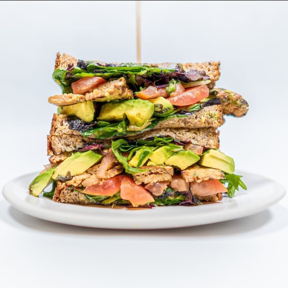 A tall, healthy sandwich with avocado, tomatoes, and greens on whole wheat bread, on a white plate.